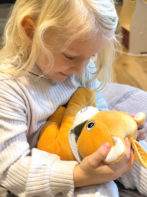 Ein Kind sitzt gemütlich und hält Lenabie Tali, den gelben Puma-Plüschcharakter, liebevoll im Arm. Das Kind blickt lächelnd nach unten auf Tali, die einen altersgerechten Lerninhalt über den eingesetzten Lenabo Smart Speaker wiedergibt. Die Szene wirkt ruhig und warm, in einer warmen Wohnatmosphäre.